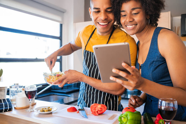 Portrait of young latin couple using a digital tablet and smiling while cooking in kitchen at home. Relationship, cook and lifestyle concept. - 写真・画像