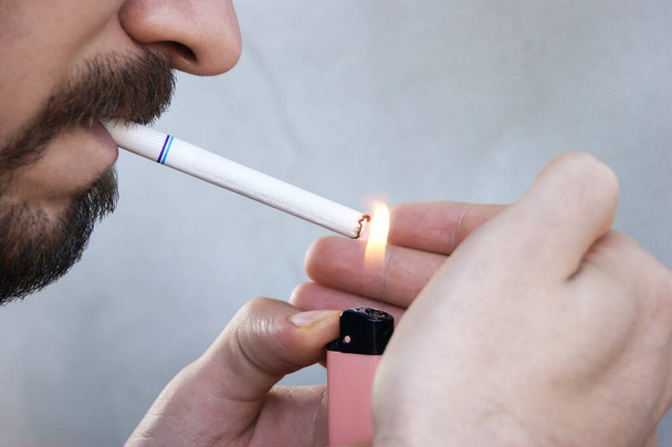 Γενειοφόρος νεαρός με γένια ανάβει τσιγάρο και καπνίζει. Κακή συνήθεια του καπνίσματος ως σημαντικό πρόβλημα της νεολαίας - Φωτογραφία, εικόνα