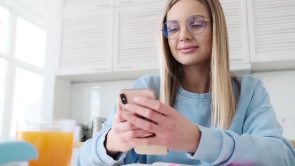 Una guapa joven rubia con gafas está usando su teléfono inteligente sentado en la cocina de su casa
 - Metraje, vídeo