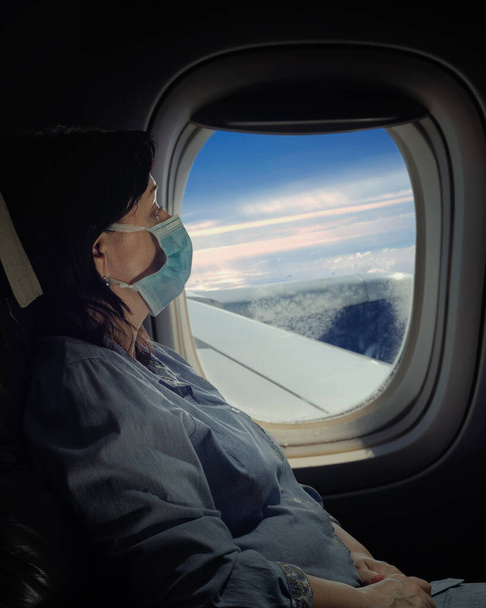 Dojrzała kobieta w masce podróżująca samolotem. Nocne ujęcie naprzeciwko okna. - Zdjęcie, obraz