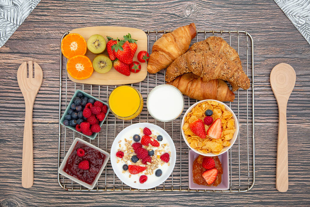 Snídaně podávaná ráno s ovocným jogurtem, máslovým croissantem a kukuřičnými vločkami Celozrnná zrna a rozinky s mlékem v šálcích a jahodami, malinami, Kiwi, čerstvým pomerančovým džusem na snídaňovém stole. - Fotografie, Obrázek