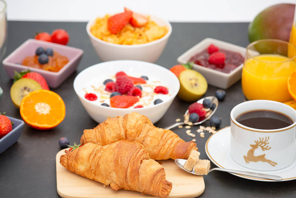 Desayuno servido por la mañana con café, croissant de mantequilla y copos de maíz Granos integrales y pasas con leche en tazas y fresa, frambuesa, kiwi, zumo de naranja fresco en la mesa de desayuno. - Foto, Imagen