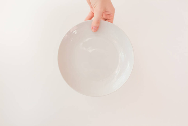  Female hand holds a white round plate on a white background as a mockup with copy space. Ceramic flat saucer. - Фото, изображение