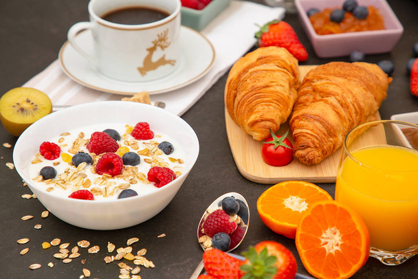 Desayuno servido por la mañana con café, croissant de mantequilla y copos de maíz Granos integrales y pasas con leche en tazas y fresa, frambuesa, kiwi, zumo de naranja fresco en la mesa de desayuno. - Foto, imagen