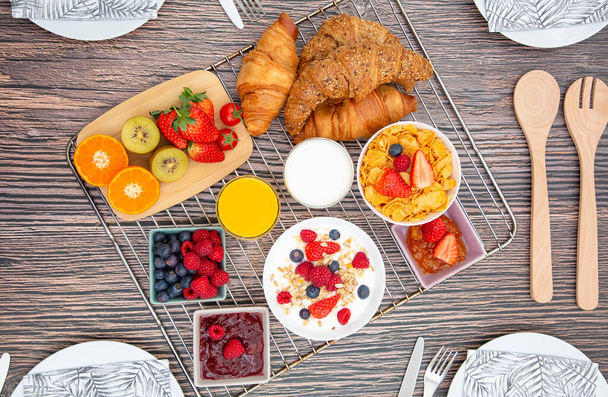 Café da manhã servido com iogurte de frutas, croissant de manteiga e flocos de milho Grãos integrais e passas com leite em xícaras e morango, framboesa, Kiwi, suco de laranja fresco na mesa de café da manhã
. - Foto, Imagem