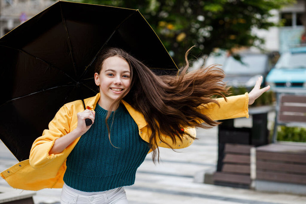 Una giovane ragazza adolescente cammina per la città con il tempo piovoso. La ragazza è vestita con vestiti colorati e stivali di gomma - Foto, immagini