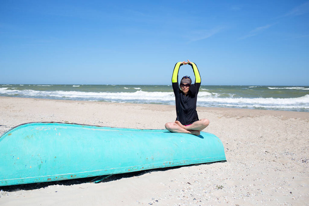 Femme mature séance d'entraînement d'exercice se détendre et méditer à la belle plage de la mer venteuse, s'asseoir dans la pose de lotus padmasana au vieux bateau bleu et lever les mains - Photo, image