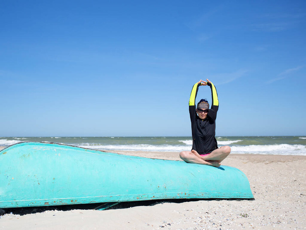 Žena ve větrovce a slunečních brýlích sedí u větrného písku mořská pláž na staré modré lodi v józe lotos padmasana póza, cvičení volný čas, sportovní aktivita během dovolené  - Fotografie, Obrázek