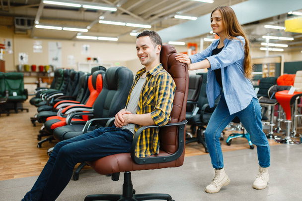 Happy young couple rides on office chair in furniture store showroom. Man and woman looking samples for bedroom in shop, husband and wife buys goods for modern home interior - Fotoğraf, Görsel