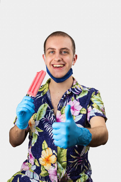 Een man die rode ijsjes eet, lacht en geniet van de zomer. Hij droeg een Hawaïaans shirt, beschermende handschoenen en gezichtsmasker op zijn kin. Coronavirus pandemische preventie tijdens de zomer - Foto, afbeelding