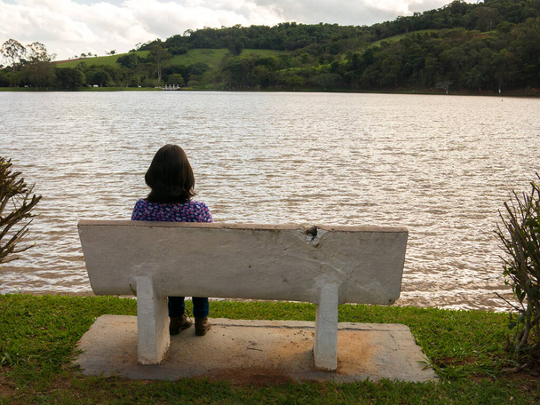 Menina não reconhecida sentada em um assento na frente de um lago na luz de fundo
 - Foto, Imagem