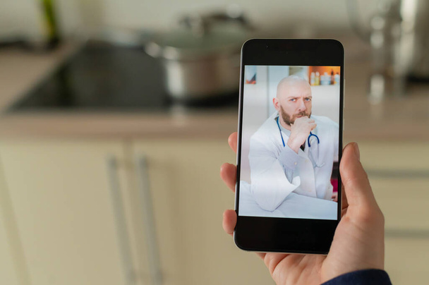 A serious bearded doctor communicates with a patient via a video call on a smartphone. Online medicine concept. Woman in the kitchen takes therapist remote consultation on mobile. - Fotó, kép