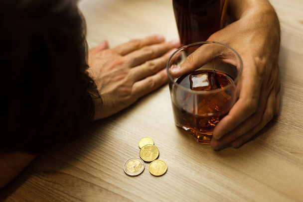Καταθλιπτικός, μεθυσμένος, κοιμάται στο τραπέζι, με ένα ποτήρι κονιάκ στο χέρι, μπουκάλι και τα τελευταία νομίσματα. έννοια της φτώχειας, του αλκοολισμού και της κατάθλιψης - Φωτογραφία, εικόνα