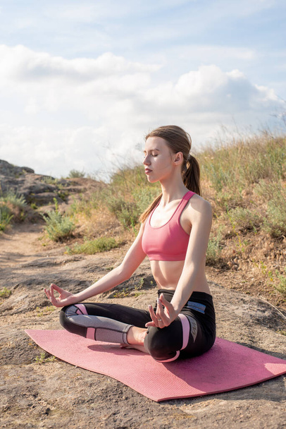 Side view of young caucasian woman meditating,breathing,practicing yoga lotus pose on mat in the sunny field,blue sky background.Girl wearing pink sportswear,relaxing with namaste.Healthy lifestyle. - Foto, Bild