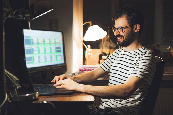 Μοντέρνος νεαρός που δουλεύει εξ αποστάσεως από το σπίτι του τη νύχτα. Casual millennial τύπος πληκτρολογώντας στον υπολογιστή notebook surfing internet κοιτάζοντας την οθόνη απολαμβάνοντας μακρινή δουλειά - Φωτογραφία, εικόνα