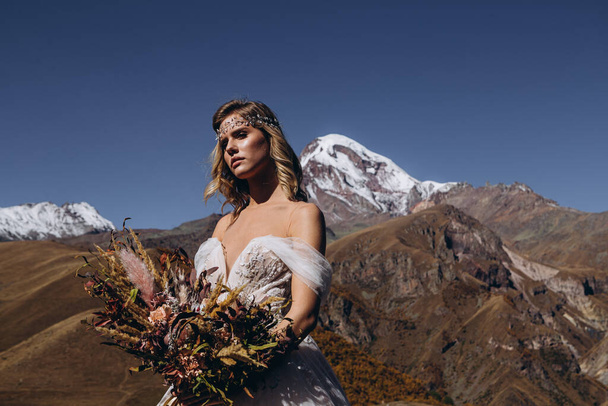 photo of a beautiful bride dressed in a long wedding dress and with a bouquet in her hands against the background of large snowy mountain peaks - Zdjęcie, obraz