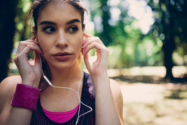 Closeup of attractive sportswoman with brown hair putting earphones and preparing for running in nature on a beautiful sunny day. - 写真・画像