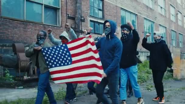 仮面をつけた顔をした男たちがアメリカ国旗を掲揚し、モットーを叫びながらスラム街を歩く。暴動で屋外に抗議する多民族男性反政府勢力. - 映像、動画
