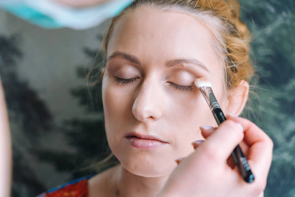 Process of making makeup. Make-up artist applied with brush on model face. Portrait of young ginger woman in beauty salon. Hand of make-up master with brush make perfect skin and color eyeshadow.  - Фото, зображення