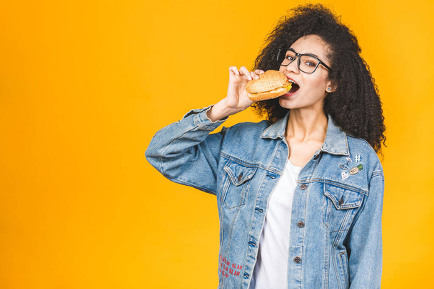 アフリカ系アメリカ人黒人の若い女性が黄色の背景で隔離されたハンバーガーを食べる. - 写真・画像