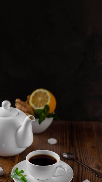 White ceramic teapot, tea set with dark brown hot drink, and natural healthy flavours lemon, lump sugar, mint and ginger on old wooden table near dark wall. Vertical banner, copy space. Rustic style. - Photo, image