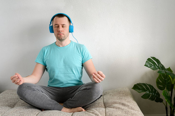 Young man sits in lotus position on the couch, listens to music on headphones. Yoga at home. - Photo, Image