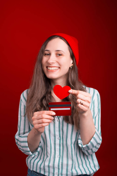 Happy smiling girl is enjoying using her credit or debit card while holding one and a small heart over red background - Foto, Imagem