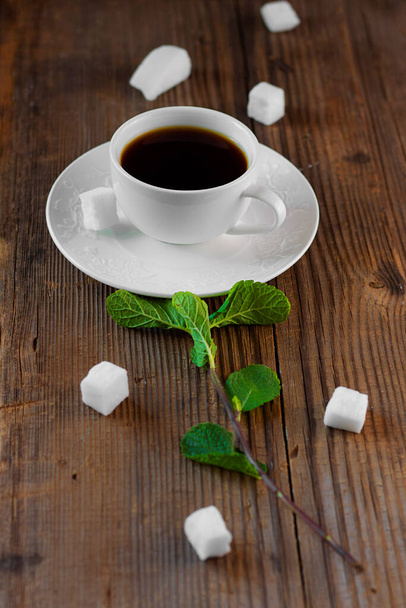 white porcelain tea-set with hot drink in on brown rustic wooden table with fresh green mint sprig put together like flower surrounded by pieces of sugar . Creative food concept. Vertical, copy space - Фото, изображение