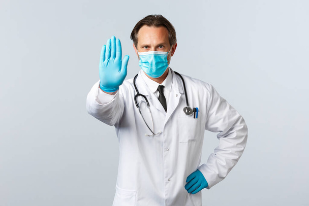 Covid-19, preventing virus, healthcare workers and vaccination concept. Serious doctor in medical mask and gloves extend hand to show stop, preventing, disagree, or restrict, demand social distancing - Photo, Image