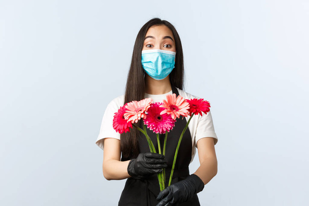 Small business, coronavirus pandemic, orders and shops concept. Enthusiastic cute asian female employee, florist store owner in medical mask and gloves give customer bouquet - Foto, Imagen