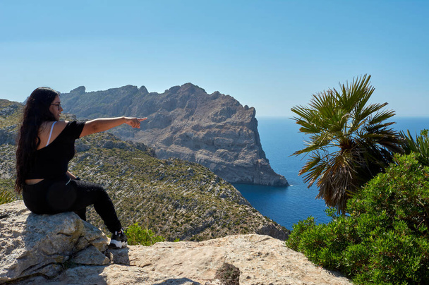 Latin girl, happy, with long brunette hair, wavy posing for, portrait, on a day out with her family in spring, sitting on the cliff of Cap de Formentor, pointing to where the lighthouse is, Mallorca, Balearic islands, Spain, - Photo, Image