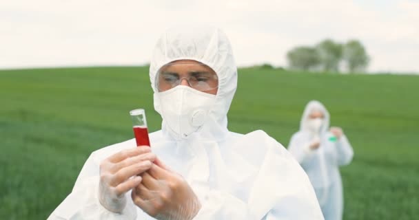 Portrait shot of Caucasian man scientist in white protective clothes and goggles holding test tube with red chemicals in green field and exploring pesticides. Woman biologist on background. - Video