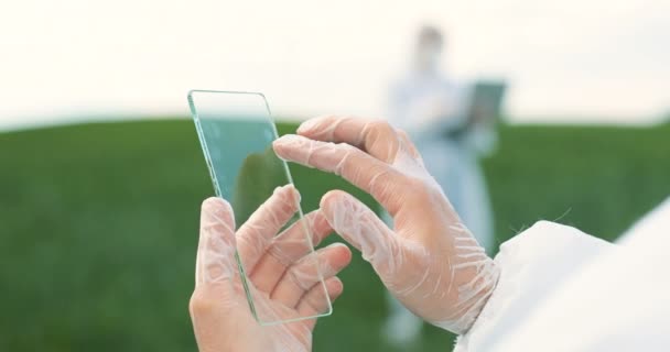 View over shoulder of farmer ecologist in protective suit tapping on glass transparent screen. Close up of futuristic device in field Touchscreen of hi-tech Scientist with smartphone of future in hand - Video