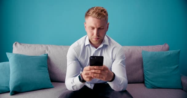 Serious man looking at phone screen while sitting on sofa - Záběry, video
