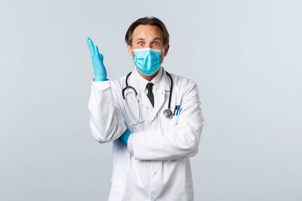 Covid-19, preventing virus, healthcare workers and vaccination concept. Excited caucasian male doctor in medical mask and gloves, raise hand up, have idea, think-up plan, white background - Photo, Image