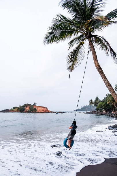 Chica de pelo azul oscuro divirtiéndose en un columpio de neumáticos de palmera en Sri Lanka. Un columpio de cuerda derecho al océano en la playa de Mirissa
. - Foto, imagen