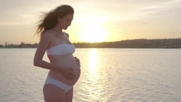Zwangere vrouw houdt haar buik wanneer staan bij zonsondergang op de zee. - Video