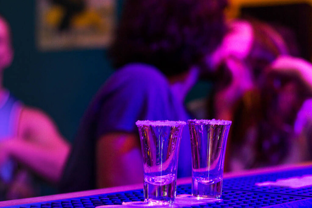 Tequila disparos en la mesa del bar en la fiesta nocturna, personas en el fondo, tragos completos alcohólicos con luz led azul en el club nocturno Tbilisi
. - Foto, imagen