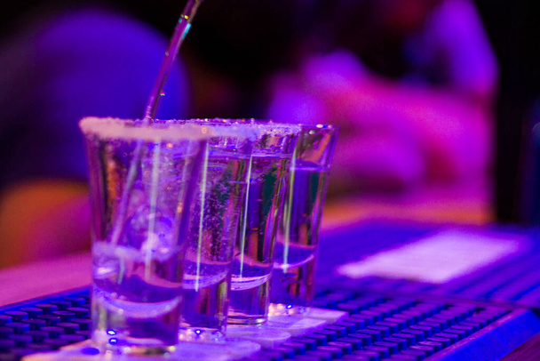 Tequila disparos en la mesa del bar en la fiesta nocturna, personas en el fondo, tragos completos alcohólicos con luz led azul en el club nocturno Tbilisi
. - Foto, imagen