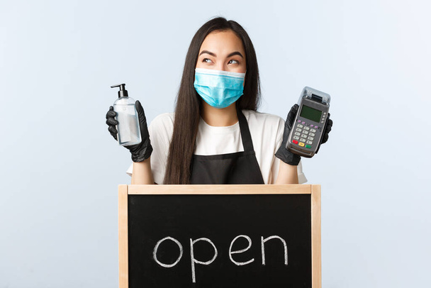 Small business, covid-19 pandemic, preventing virus and contactless orders concept. Excited cute asian waitress, barista in medical mask giggle near open sign, hold hand sanitizer and POS terminal - Photo, Image