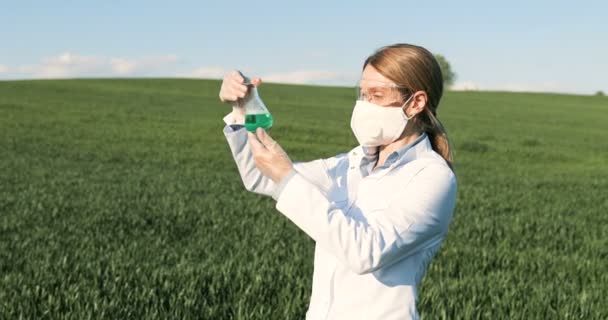 Καυκάσια όμορφη γυναίκα οικολόγος επιστήμονας σε λευκό φόρεμα, μάσκα και googles στέκεται στο πράσινο πεδίο και κοιτάζοντας χημικές ουσίες σε δοκιμαστικό σωλήνα. Γυναίκα ερευνήτρια και βιολόγος στο περιθώριο που μελετά φυτοφάρμακα. - Πλάνα, βίντεο