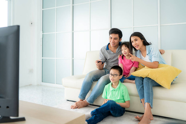 Aziatisch gelukkig gezin met vader, moeder, zoon en dochter zitten op de bank en televisie kijken thuis concept van familie waarden, vakantie dag, vakantie, geluk of levensstijl. - Foto, afbeelding