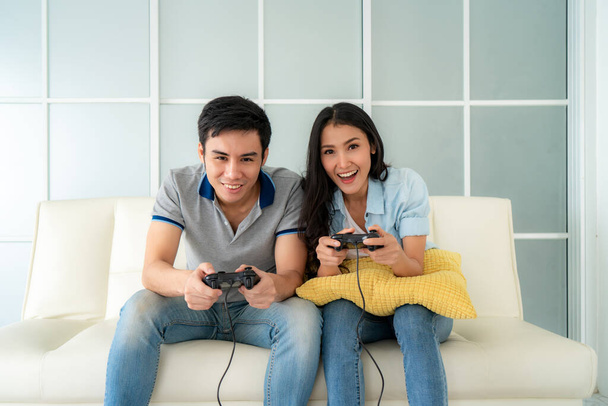 アジアのカップルの男性は、家庭でソファに座っている間にジョイスティックでビデオゲームをプレイする女性を意味し、家族の価値観と現代のゲームの概念. - 写真・画像