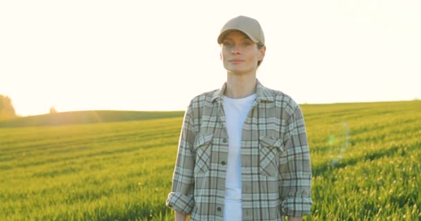 Portrait shot of beautiful young Caucasian woman in hat standing in green field and smiling cheerfully to camera. Female farmer looking straight with smile outdoor in summer. - Filmmaterial, Video