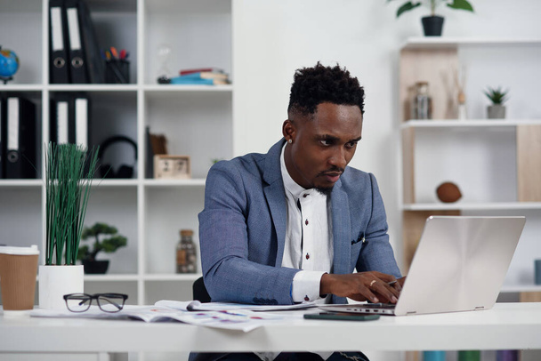 Businessman with laptop. Young African businessman is typing something on laptop in his office. - 写真・画像