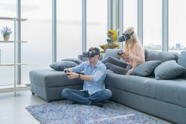 Ένα δυτικό νεαρό ζευγάρι που φοράει γυαλιά εικονικής πραγματικότητας ή γυαλιά εικονικής πραγματικότητας, ακουστικά που κάθονται και παίζουν ένα βιντεοπαιχνίδι στον καναπέ μπροστά από την τηλεόραση στο σπίτι στην έννοια της τεχνολογίας και της καινοτομίας. Διασκεδάζω. Διασκέδαση - Φωτογραφία, εικόνα
