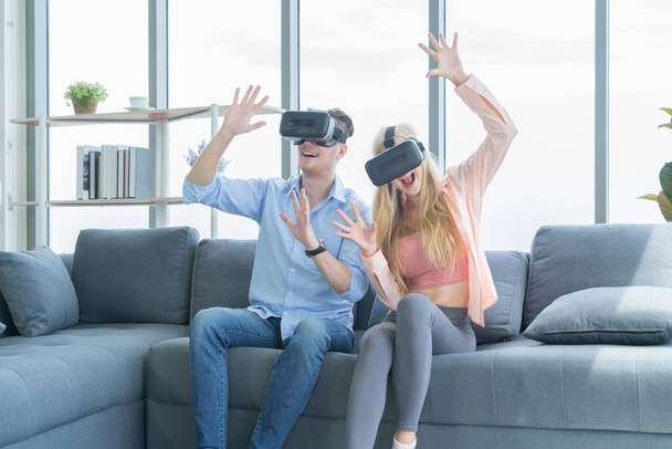 Una giovane coppia occidentale che indossa occhiali VR o realtà virtuale, cuffie sedute e che gioca a un videogioco sul divano davanti alla TV a casa nel concetto di tecnologia e innovazione. Mi diverto. Spettacolo
 - Foto, immagini