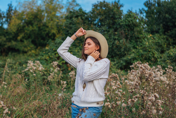 Bella ragazza bruna adolescente vestita con un maglione bianco, jeans e cappello di paglia da cowboy in piedi in erba secca marrone bur, sorridente e ridente durante il tramonto - Foto, immagini