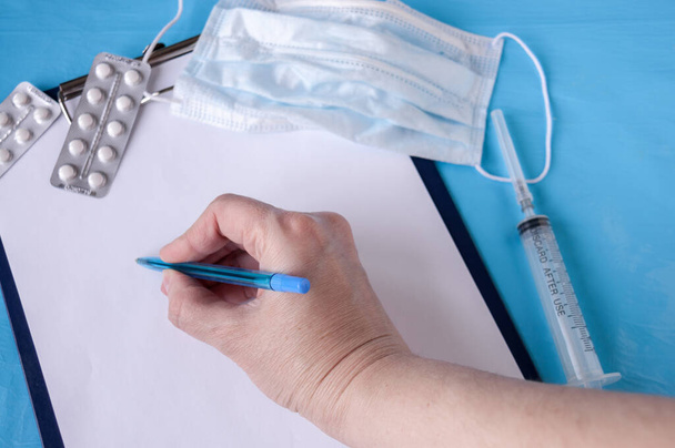 Hand writing pen on a tablet for prescriptions, medical mask, tablets, syringe on a blue background - 写真・画像
