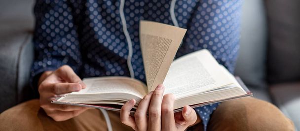 Άνδρας φοιτητής χέρι άνοιγμα σελίδα βιβλίο ανάγνωση βιβλίο στον καναπέ στη βιβλιοθήκη. Εκπαιδευτική έρευνα και αυτομελέτη. Παγκόσμια Ημέρα Βιβλίου - Φωτογραφία, εικόνα